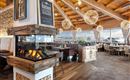 Ein gemütliches Restaurant mit Holzdecke und modernen Lampen. Im Vordergrund befindet sich ein Holzofen, während große Fenster einen Blick auf die Berge bieten.