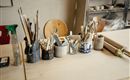 Eine Werkstatt mit verschiedenen Werkzeugen und Pinsel in Behältern. Der Tisch ist aus leichtem Holz und im Hintergrund sind weitere Materialien sichtbar.