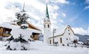 Ein verschneites Dorf mit einer schönen Kirche und einem hohen, grünen Kirchturm. Die Umgebung ist ruhig und winterlich, bedeckt mit frischem Schnee.