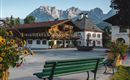 A picturesque village square with traditional wooden houses and blooming flowers. In the background, majestic mountains rise under a clear sky.