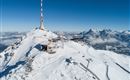 Ein schneebedeckter Berggipfel mit einem Sendemast und einer Berghütte. Im Hintergrund sind majestätische Berge und ein klarer blauer Himmel sichtbar.