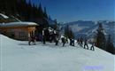Eine winterliche Landschaft mit viel Schnee und Skifahrern an einer Liftstation. Im Hintergrund sind schneebedeckte Berge und Tannenbäume zu sehen.