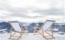 Two lounge chairs are on snow-covered ground overlooking snow-capped mountains. The sky is cloudy and the landscape appears calm and peaceful.