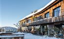 A modern wooden building in the snow with large windows and a clear blue sky. The surroundings are wintry and inviting.