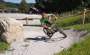 Ein Radfahrer fährt schnell auf einer kurvigen Strecke. Die Umgebung ist grün, mit einem Felsen und Bäumen im Hintergrund.