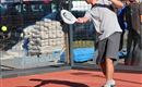 A man is playing padel tennis on a red court. The sun is shining and construction work can be seen in the background.