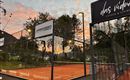 A tennis court with a bright orange surface is surrounded by a fence. In the background, a beautiful sunset and some trees can be seen.