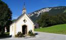 Eine kleine Kapelle steht an einer Weggabelung, umgeben von grünen Wiesen und Bäumen. Im Hintergrund sind majestätische Berge sichtbar unter einem klaren blauen Himmel.