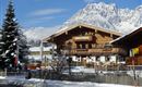 Ein schönes Holzhaus im Winter mit schneebedecktem Dach und malerischer Berglandschaft im Hintergrund. Der Himmel ist klar und blau.