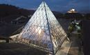 Eine moderne Glaspyramide leuchtet in der Dämmerung. Im Hintergrund ist eine Burg auf einem Hügel sichtbar.