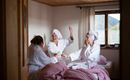 Three women in bathrobes and with towels on their heads are sitting in a cozy room. They seem to be laughing and enjoying time together.