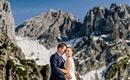 A bride and groom pose in front of an impressive mountain landscape. The peaks are snow-covered and the sky is clear.