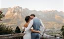 A bride and groom are standing on a porch and embracing each other. In the background, majestic mountains and a picturesque landscape can be seen.