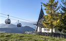 Eine malerische Berglandschaft mit einer Kapelle und Seilbahnkabinen. Im Hintergrund sind sanfte Hügel und die Alpen sichtbar.