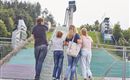 Eine Familie steht vor einer Sprungschanze im Freien. Im Hintergrund sind grüne Hügel und Sitzplätze zu sehen.