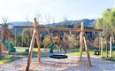A playground with a wooden swing and various play equipment. In the background, there are trees and hills.
