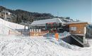 A modern ski chalet in a snowy mountainous area. In the background, ski lifts and snow-covered trees can be seen.