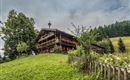 A traditional wooden house surrounded by green meadows and trees. In the background, gentle hills and a cloudy sky can be seen.