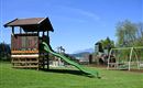A playground with a wooden slide and swings outdoors. In the background, green meadows and trees can be seen.