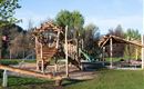 A large wooden playground with various climbing elements and a slide. In the background, there are trees and a meadow.