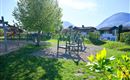 A playground in a green environment with a large tree. In the background, mountains and houses can be seen.