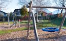 A playground with various play equipment, including a swing and a slide. The surroundings are green with trees and a quiet residential house in the background.