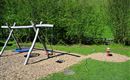 A playground with a wooden swing and a round sand area. In the background, green bushes can be seen.