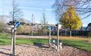 A playground with a wooden climbing structure and a bench. In the background, there are trees and some buildings visible.