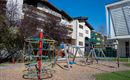 Ein Spielplatz mit einer Kletterstruktur und Schaukeln. Im Hintergrund sind mehrere Wohngebäude zu sehen.