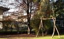 A playground with a wooden swing is located in a garden. In the background, trees and a house are visible.