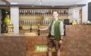 Een vriendelijke man in traditionele Beierse outfit staat achter de bar. Achter hem staan talrijke tapkranen voor bier opgesteld.