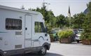 A motorhome in a parking lot, surrounded by trees and plants. In the background, a church steeple is visible.