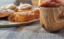 A handcrafted wooden cup is held by a hand. In the background, various pastries are visible on a plate.