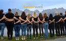 A group of people stands with their backs to the camera and looks at the mountains. They are wearing black t-shirts and have their hands on their hips.