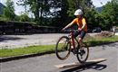 Ein Radfahrer springt über ein Holzbrett auf einer Straße. Er trägt einen Helm und ein orangefarbenes Shirt.