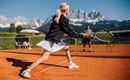 Eine Frau spielt Tennis auf einem Platz mit Blick auf beeindruckende Berge. Es ist sonnig und die Atmosphäre ist aktiv und sportlich.