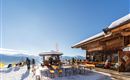 A cozy mountain restaurant with outdoor seating in the snow. The sky is blue and the surroundings are wintry and sunny.