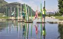 Vijf kinderen staan op platforms in het water en balanceren met kleurrijke stokken. Op de achtergrond zijn groene heuvels en een heldere lucht te zien.