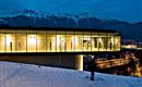 Ein modernes Gebäude mit großen Glasfenstern steht auf einem Hügel im Schnee. Im Hintergrund sind schneebedeckte Berge und ein Nachtblick auf die Stadt zu sehen.