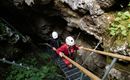 Two people are descending a staircase into a cave. They are wearing helmets and are surrounded by rocks and plants.