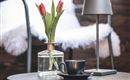 Ein stilvoller Tisch mit einer Tasse Kaffee und einer Glasvase mit frischen Tulpen. Im Hintergrund steht eine moderne Lampe und ein gemütlicher Stuhl.