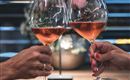 Zwei Gläser mit Roséwein werden angestoßen. Im Hintergrund sind verschwommene Blumen und sanftes Licht zu sehen.