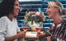 Zwei Frauen lachen und stoßen mit Weingläsern an. Im Hintergrund sind Blumen und eine moderne Umgebung zu sehen.