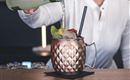 Ein Barkeeper gießt ein Getränk in einen eleganten, kupfernen Becher. Der Becher ist mit Eis, Limetten und roten Beeren dekoriert.