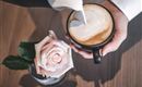 Eine Tasse Kaffee wird mit cremigem Milchschaum gefüllt. Neben der Tasse steht eine zarte rosa Rose.
