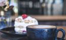 Eine Tasse Kaffee auf einem Teller neben einem Stück Kuchen. Im Hintergrund sind stilvolle Dekorationen zu sehen.