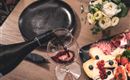 Ein Glas Rotwein wird eingeschenkt, während ein rustikales Essen auf einem Tisch präsentiert wird. Dazu gehören Käse, Wurst, Obst und frische Blumen.
