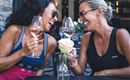 Zwei Frauen lachen und stoßen mit Gläsern an, während sie am Tisch sitzen. Auf dem Tisch steht eine Rose und einige Menükarten.