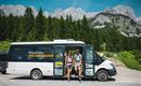 A hiking bus stands in nature, surrounded by mountains and forests. Two people get out, ready for an adventure.