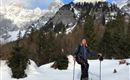 Eine Frau beim Schneeschuhwandern in einer verschneiten Berglandschaft. Im Hintergrund sind majestätische Berge und Nadelbäume zu sehen.